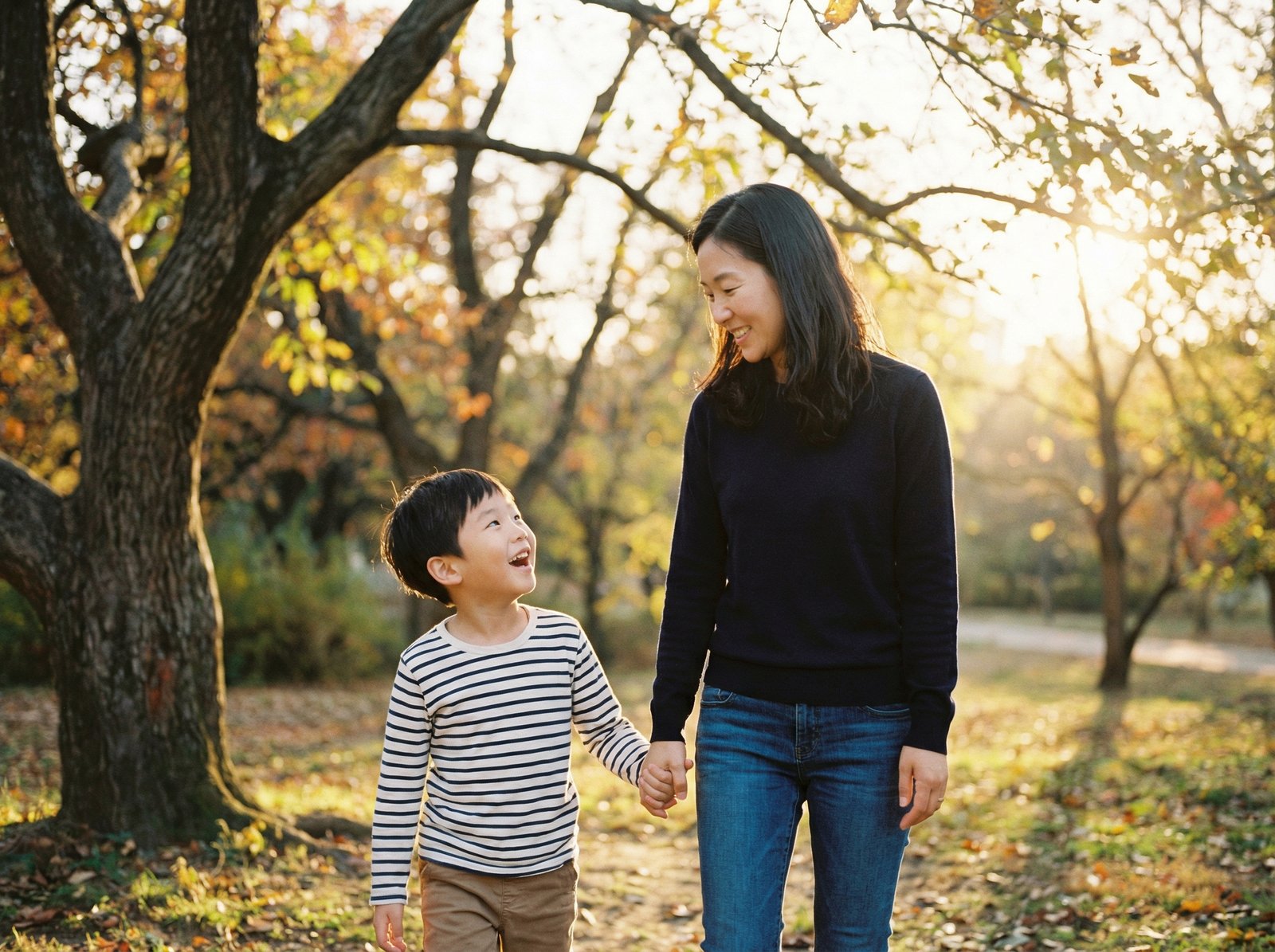 A Korean parent and a child, maybe 6-8 years old, walking hand in hand in a park, with warm sunlight. The child is looking up at the parent with a happy expression. Lifestyle photography, warm lighting, natural setting, aspect ratio 4:3, no visible text.