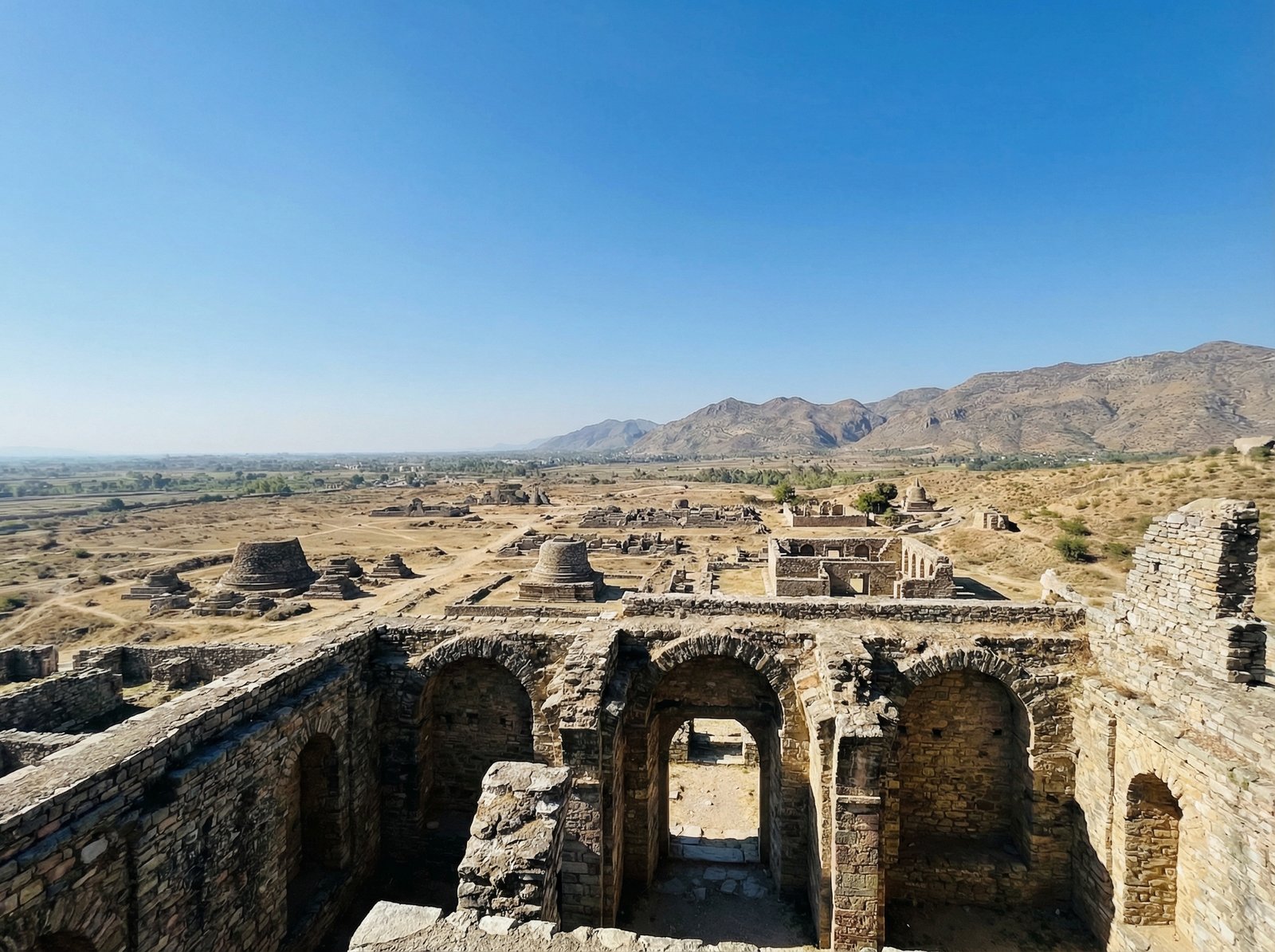 A scenic view of the ancient city of Taxila from the Julian Monastery ruins, showing a vast expanse of arid landscape with scattered ancient structures and distant mountains under a clear blue sky. Aspect ratio 4:3, no visible text.