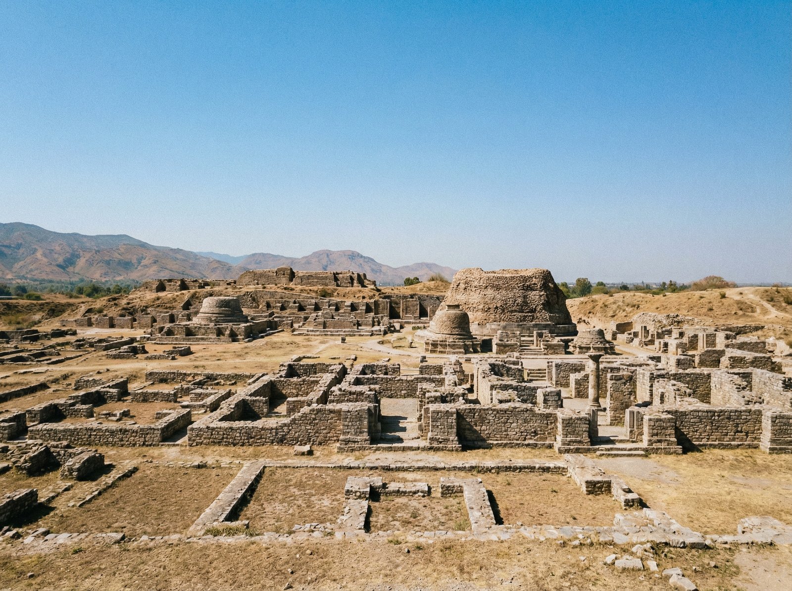 Panoramic view of the ancient ruins of Taxila, Pakistan, under a clear sky. The ruins show well-preserved stone structures and remnants of a large historical city. The landscape is dry and arid with distant hills. Aspect ratio 4:3, no visible text.