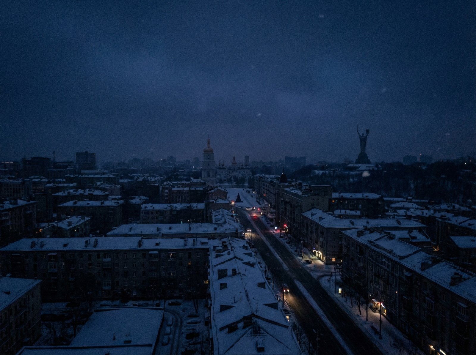 A cinematic night view of Kyiv city covered in snow with most building lights turned off due to a blackout, faint silhouettes of architectural landmarks, cold blue atmosphere, realistic photography, 4:3 aspect ratio, no text