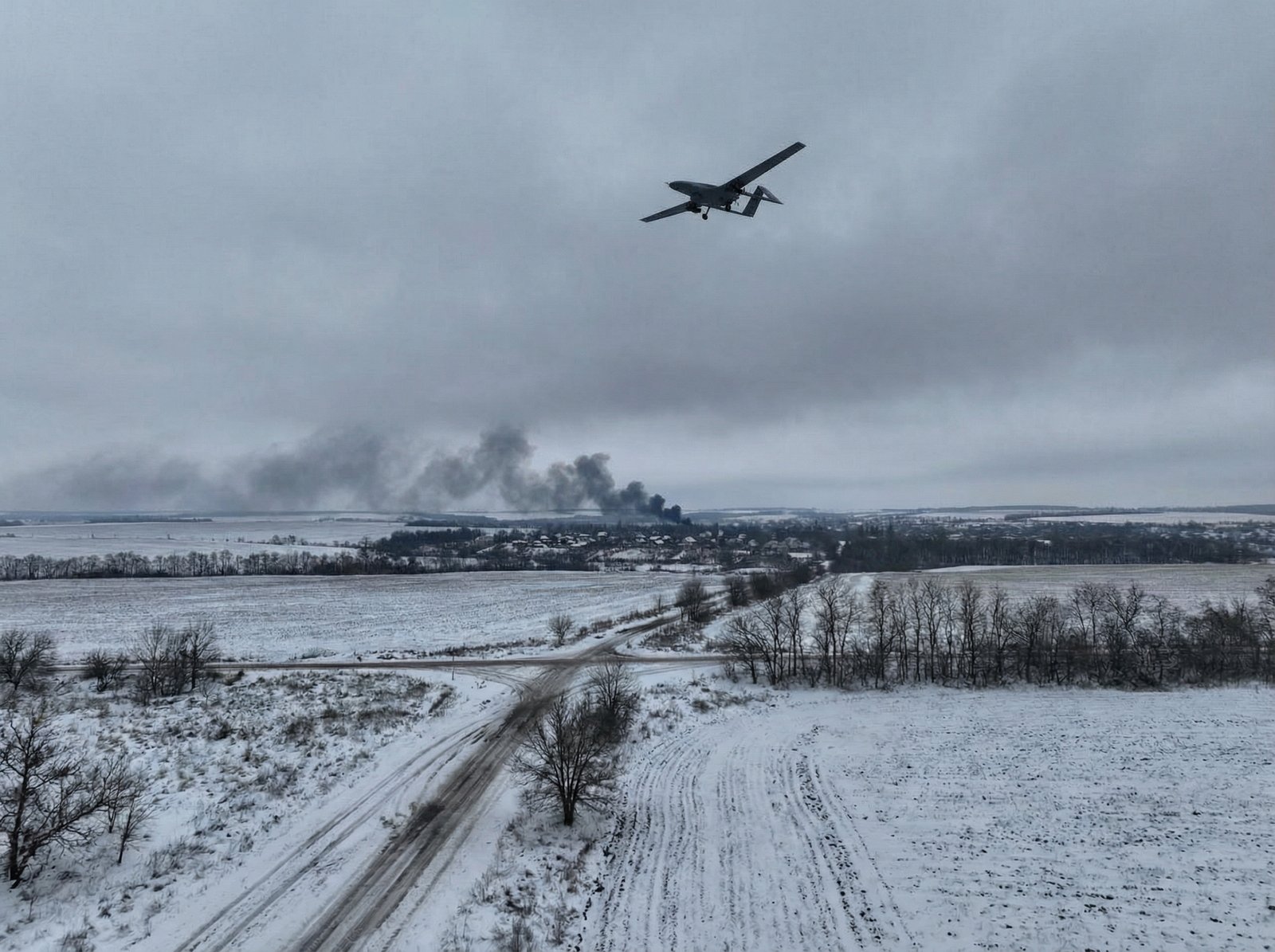 A wide shot of a snowy winter landscape in Ukraine with a military drone flying in the gray sky, smoke rising in the far distance, realistic military photography style, 4:3 aspect ratio, no text