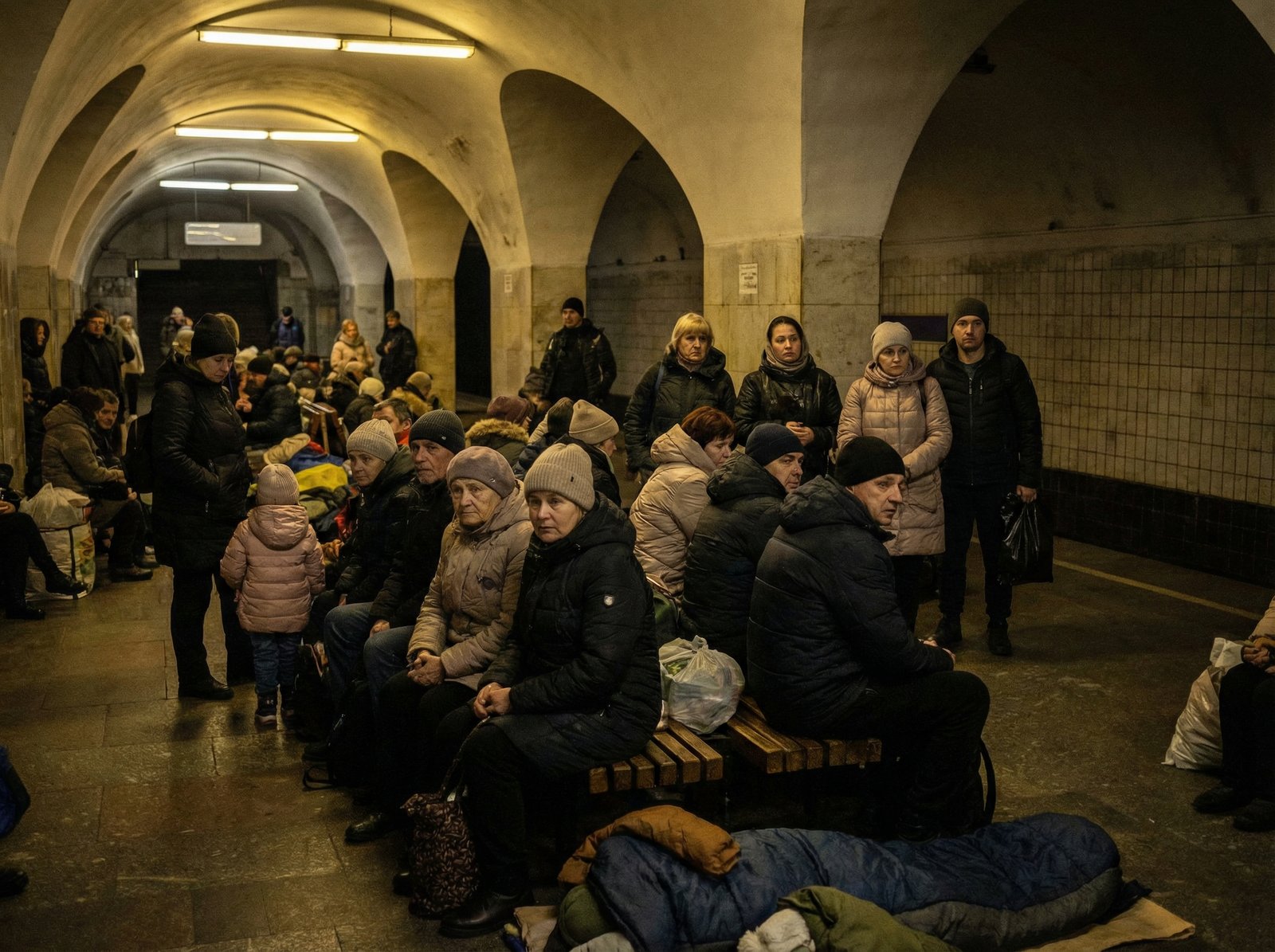 A realistic photo of many Ukrainian people taking shelter inside a deep underground metro station in Kyiv during an air raid, dim lighting, wearing winter clothes, natural expressions of anxiety and fatigue, cinematic composition, 4:3 aspect ratio, no text