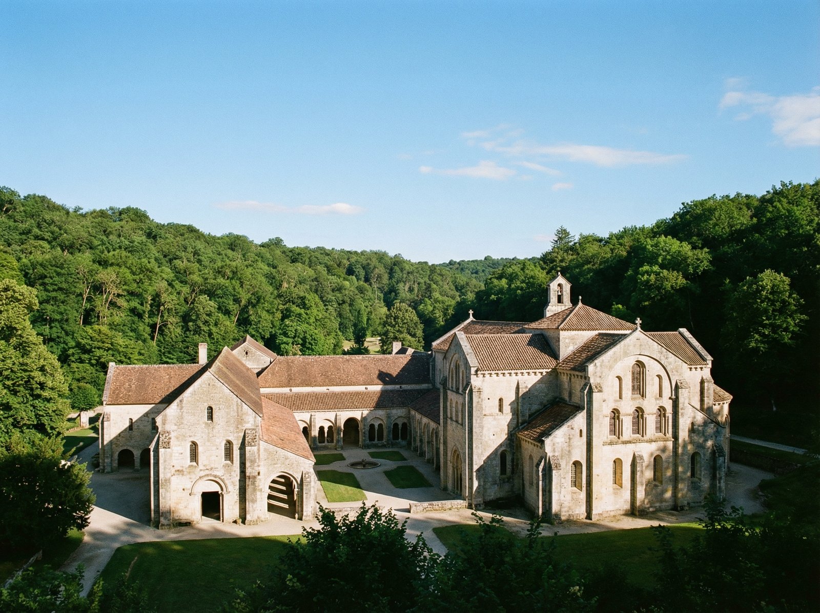 A panoramic view of the Cistercian Abbey of Fontenay in France, surrounded by lush green trees and a clear blue sky. The ancient stone buildings show intricate architectural details. No visible text. Aspect ratio 4:3.