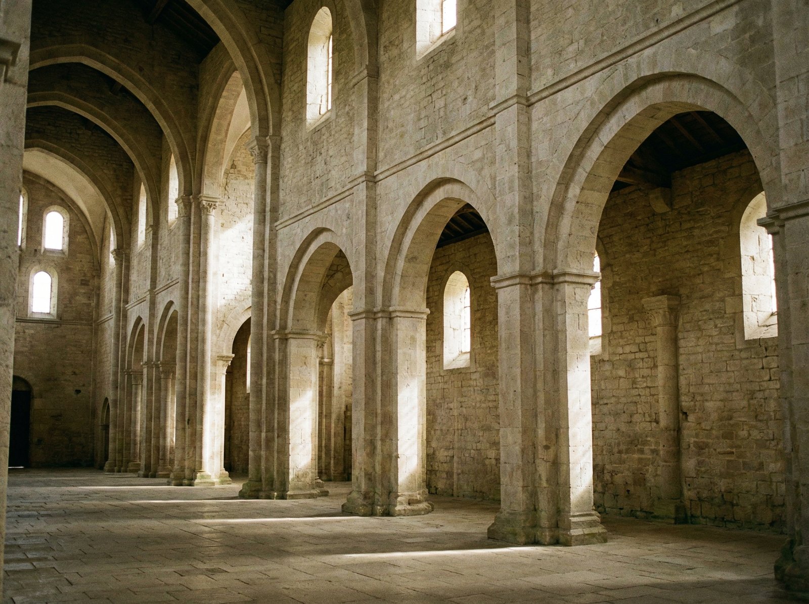 The interior of the Cistercian Abbey of Fontenay, featuring tall stone arches and simple, unadorned walls. Natural light streams in through small windows. No visible text. Aspect ratio 4:3.
