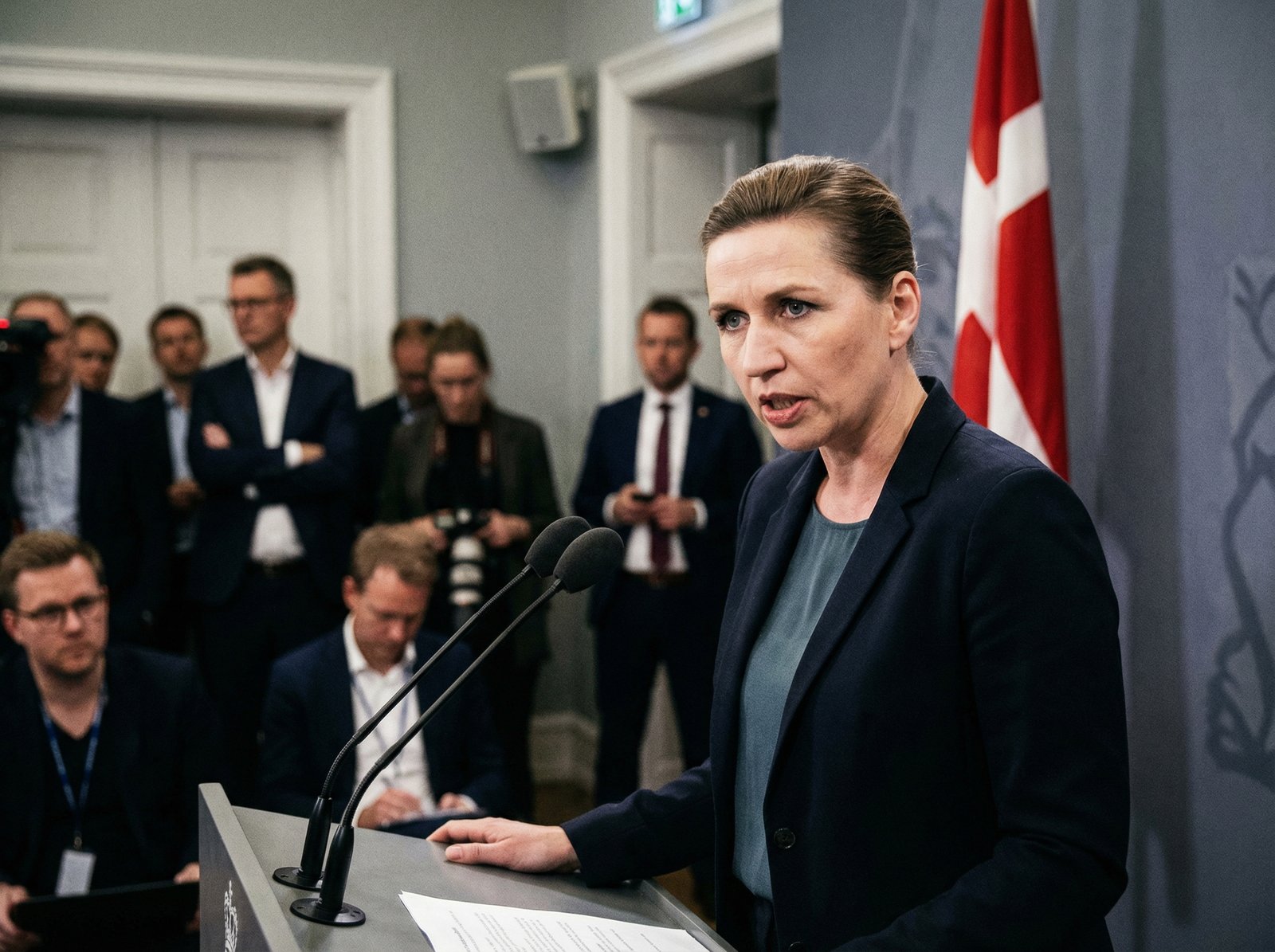 A serious Danish Prime Minister Mette Frederiksen delivering a strong warning at a press conference, with the Danish flag subtly in the background. Informational style, 4:3 aspect ratio, no visible text.