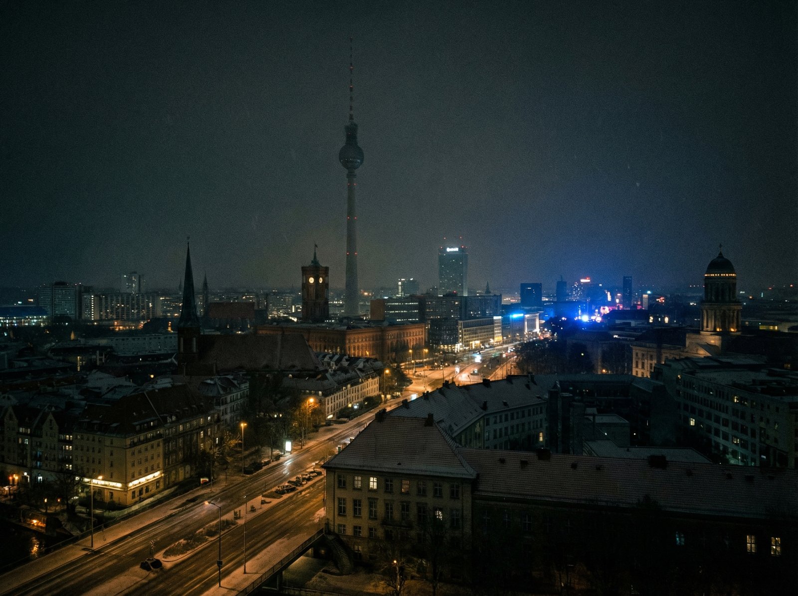 Realistic photography of Berlin city skyline at night during a massive winter power outage, few streetlights visible, snow falling lightly, emergency vehicle blue lights in the distance, cinematic atmosphere, 4:3