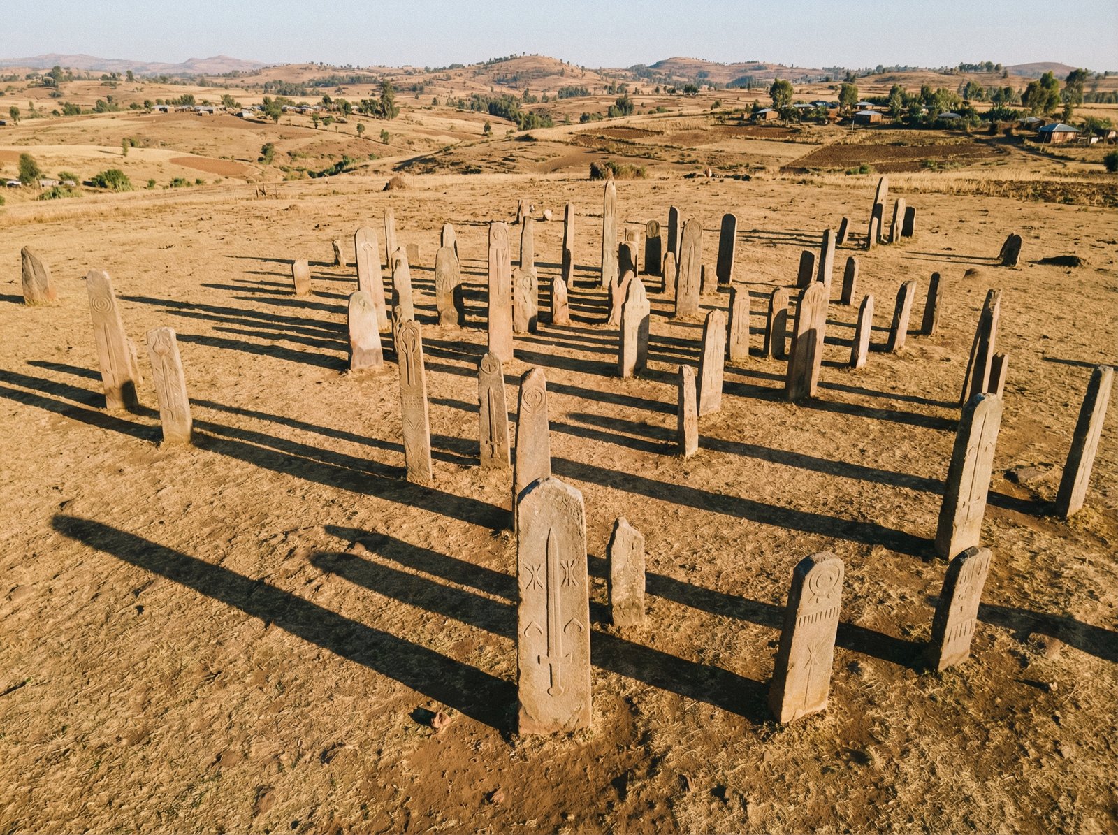 An aerial view of the Tiya archaeological site in Ethiopia, showing numerous ancient stelae standing upright in a field. The sun is shining, casting long shadows. Aspect ratio 4:3, no visible text.