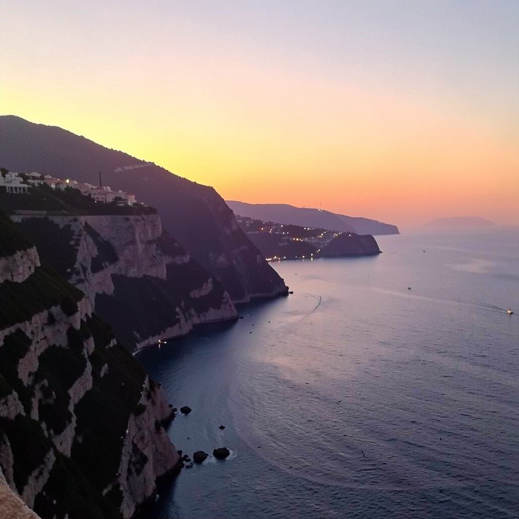 Soft sunset over the Amalfi Coast with cliffs, sea, and distant village lights, no visible text, no logos, no watermark, aspect ratio 1:1